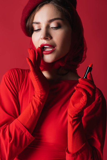 pretty woman in gloves, headscarf and beret holding lipstick isolated on red - 写真・画像