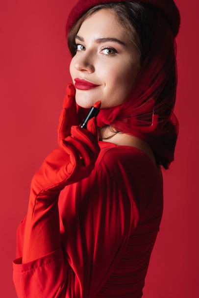 pleased young woman in gloves, headscarf and beret holding lipstick isolated on red - Photo, Image