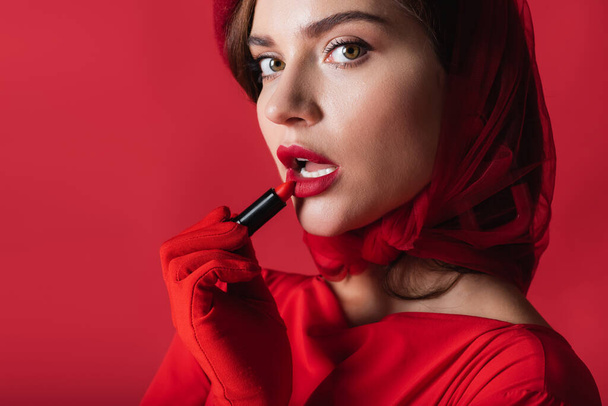 young woman in glove and beret applying lipstick isolated on red - Photo, Image