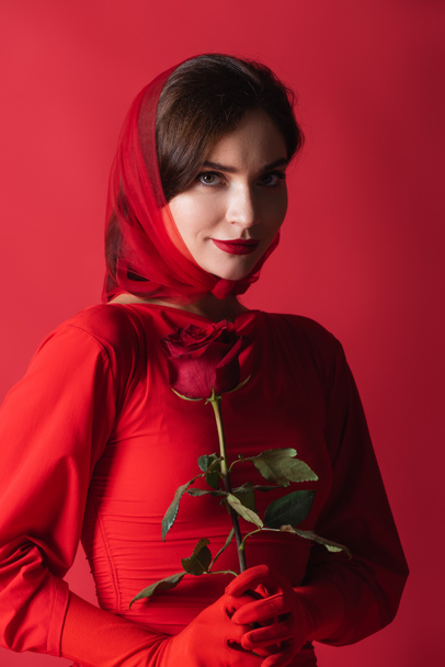 pleased young woman in gloves, headscarf and dress holding rose isolated on red - Valokuva, kuva