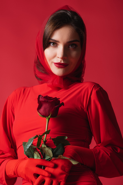young elegant woman in dress and headscarf holding rose while looking at camera isolated on red - Фото, изображение