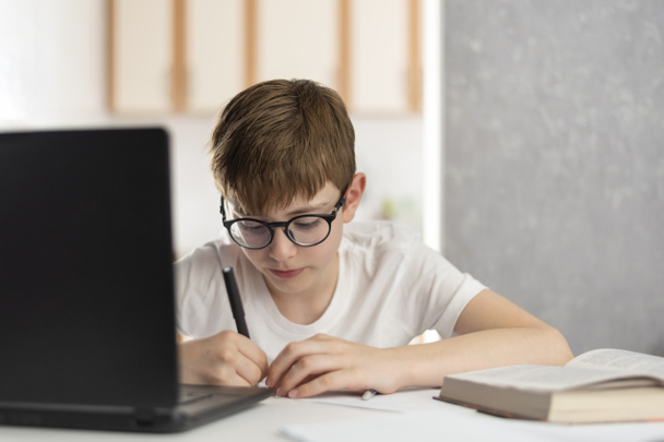 Child at home near laptop during distance learning. Online lessons. Teen boy does homework at computer. Home schooling. - Photo, Image