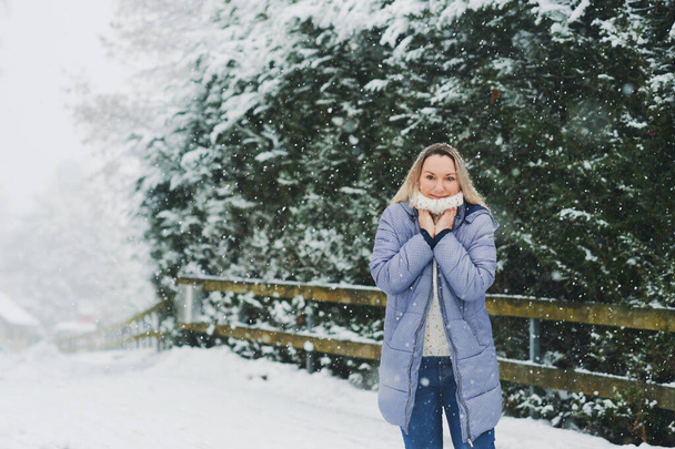 Mujer madura feliz disfrutando de un bonito día de invierno, vistiendo un cálido jersey blanco y una chaqueta azul de invierno, de pie bajo la nieve - Foto, Imagen