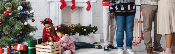 africké americké děti hrající si s cetkami v blízkosti vánočního stromku a rodiny v obývacím pokoji, banner - Fotografie, Obrázek