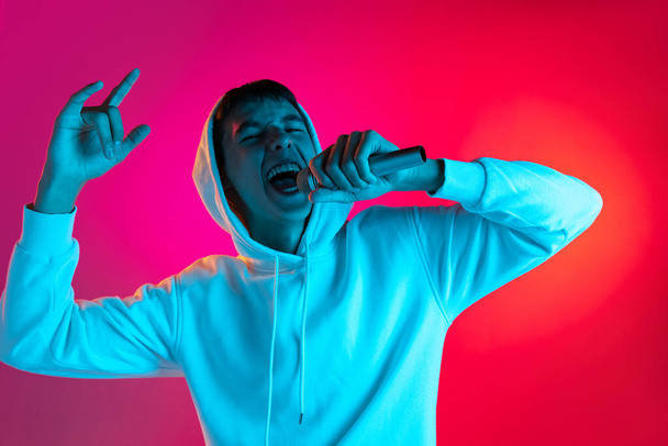 Portret van een jonge man, student of jongen in casual kleding zingen met microfoon geïsoleerd op magenta studio backgroud in neon. - Foto, afbeelding
