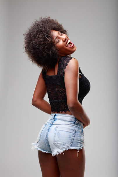 Mujer negra sexy provocativa descarada en pantalones cortos de verano que miran hacia atrás sobre su hombro y se ríen de la cámara sobre un fondo gris - Foto, Imagen