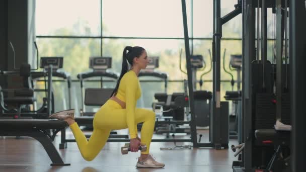 Femme sportive fait des fentes pour les jambes avec des haltères dans ses mains dans la salle de gym. Jeune femme hispanique musclée faisant de l'exercice avec des haltères dans la salle de gym En vêtements de sport jaunes - Séquence, vidéo