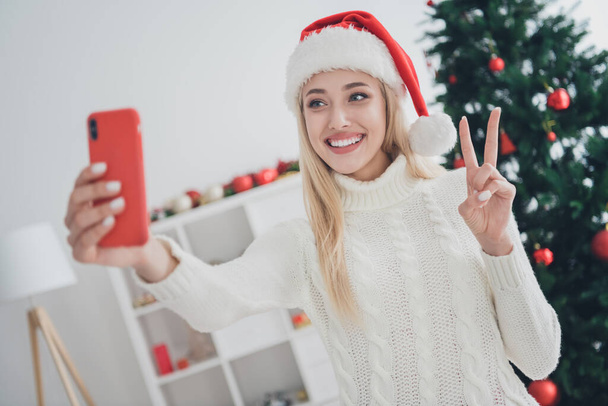 Photo of charming pretty young woman dressed white sweater recording video modern gadget showing v-sign indoors room home house - 写真・画像