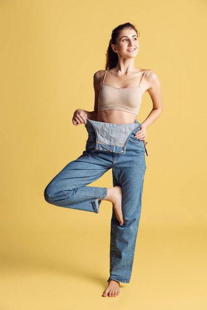 Portrait of young beautiful slim woman in blue jeans jumpersuit posing isolated over yellow studio background. Weight Loss concept. - Φωτογραφία, εικόνα