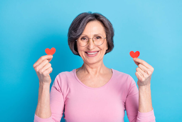 Photo of happy excited beautiful pretty mature woman in glasses hold little red paper hearts isolated on blue color background - Foto, Imagem