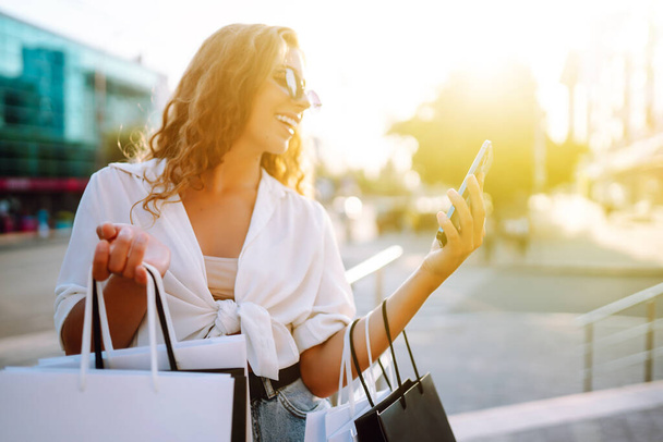 Young woman using phone, holding shopper bags, standing outdoors, choosing and ordering clothes and presents. Online shopping concept. Consumerism And Technology. - 写真・画像