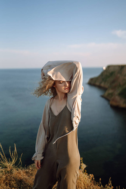 Curly woman posing at the edge of a rock above the sea, with blue sky and sea background. Autumn style. Travel and adventure. - 写真・画像