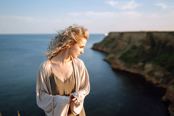Curly woman posing at the edge of a rock above the sea, with blue sky and sea background. Autumn style. Travel and adventure. - Foto, imagen