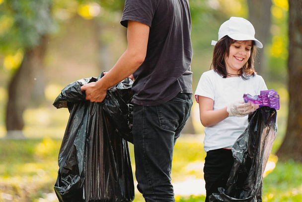 Picture of a smiling beautiful girl in protective gloves and adult man volunteer gathering plastic rubbish together outdoors using big black garbage bags. - Foto, Imagem
