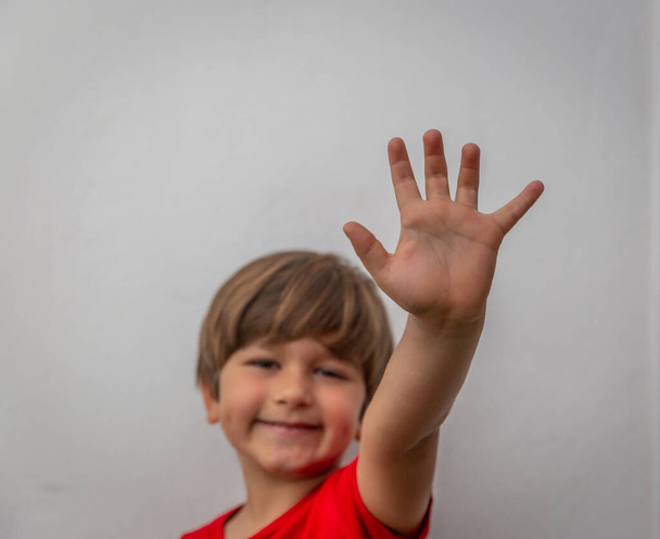 Усміхнений хлопчик у червоній футболці показує ліву руку з білою стіною на задньому плані
 - Фото, зображення