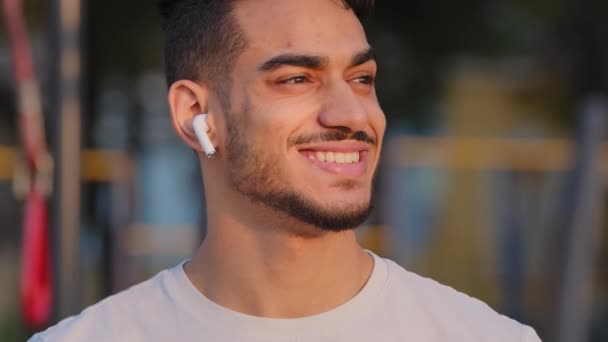 Посміхнений чоловік з Близького Сходу в сучасних навушниках, який дивиться убік на міський стадіон, щасливий індійський хлопець з білими зубами посміхається слухати музику або подкаст насолоджується хорошим звучанням в навушниках. - Кадри, відео
