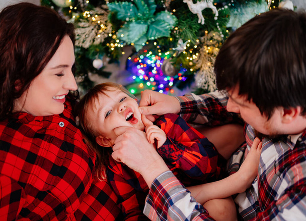 dad, mom and little daughter in red plaid pajamas playing at the Christmas tree - Photo, image