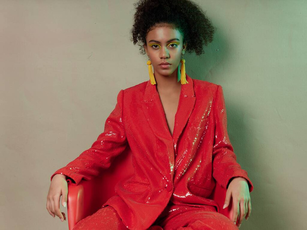 mulatto woman in a red jacket and trousers sits on a chair indoors hairstyle earrings - Fotografie, Obrázek