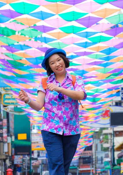 Szczęśliwa azjatycka turystka w luźnej odzieży uśmiechnięta wesoło podczas spaceru ulicą z barwną dekoracyjną tkaniną wiszącą na powietrzu podczas letniego festiwalu wakacyjnego - Zdjęcie, obraz