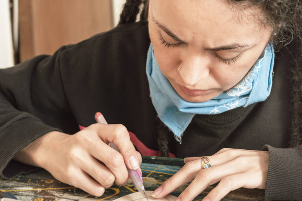 pretty young white girl Hispanic latina art director, with dreadlocks and a frown on her face, sitting at her work table cutting cardboard in great detail with a cutter, in her workshop. - Photo, Image