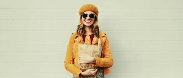 Portrait of beautiful smiling young woman holding grocery shopping paper bag with long white bread baguette on gray background - Fotografie, Obrázek