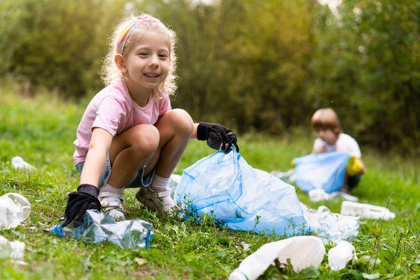 τα παιδιά αφαιρούν πλαστικά σκουπίδια και τα βάζουν σε μια βιοδιασπώμενη σακούλα σκουπιδιών στην ύπαιθρο. Η έννοια της οικολογίας, της επεξεργασίας αποβλήτων και της προστασίας της φύσης. Προστασία του περιβάλλοντος. - Φωτογραφία, εικόνα