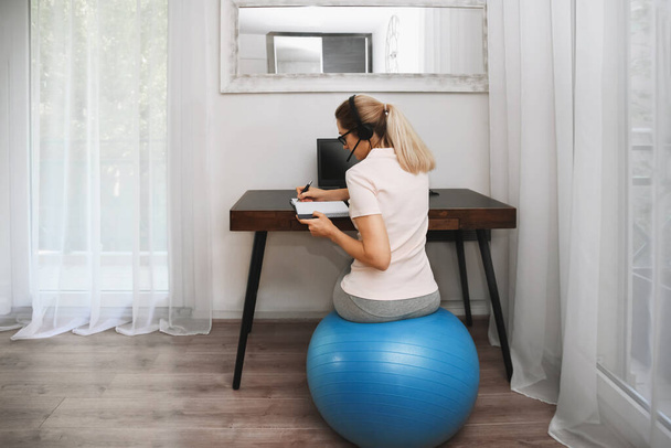A woman sits on a fitness ball in headphones and works at home during quarantine - Φωτογραφία, εικόνα