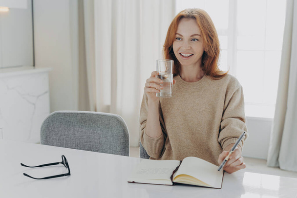 Feliz mujer pelirroja con vaso de agua mineral pura escribiendo lista de compras en cuaderno en casa - Foto, imagen
