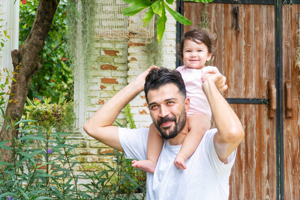 Cheerful father holding and piggyback his baby daughter in his neck while relax time at home. Enjoyment little lovely mixed race girl over daddy shoulder in the garden . Happy fathers day concept. - Photo, Image