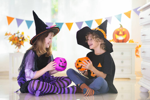 Dziewczynka i chłopiec w stroju czarownicy na Halloween cukierek lub psikus. Dzieciaki trzymające cukierki w wiadrze z dyniami. Dzieci świętują Halloween w urządzonym kominku. Sztuczka rodzinna lub psikus. - Zdjęcie, obraz