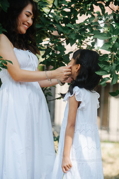 Happy mother pinching her daughter's cheeks, they stand under a short tree in a park, looking at each other with love and adoration. They both wearing white clothes. - Fotoğraf, Görsel