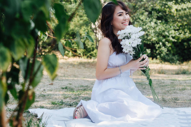 Smiling asian woman sitting on a blanket in a park, holding bouquet of white flowers She's wearing white dress, having picnic in a park, enjoying last warm days of the early fall in tree shadow. - 写真・画像