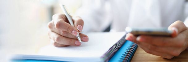 Γυναίκα χέρι κρατά smartphone στο χέρι της και κρατά σημειώσεις στο σημειωματάριο - Φωτογραφία, εικόνα