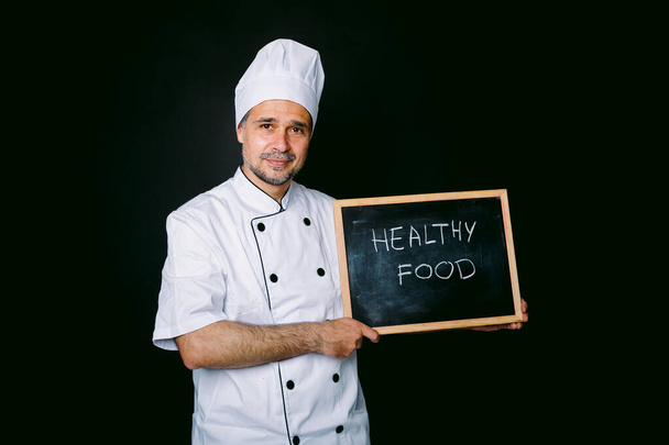 白いジャケットと帽子に身を包んだ料理は、黒の背景に「ビーガン」と書かれた黒板を持っています。健康食品とレストランのコンセプト. - 写真・画像