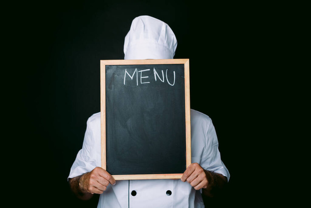 Un chef vêtu d'un chapeau blanc et d'une veste recouvre son visage d'un tableau noir qui se lit comme suit : Menu sur fond noir. Restaurant, cuisine et concept d'école de cuisine. - Photo, image