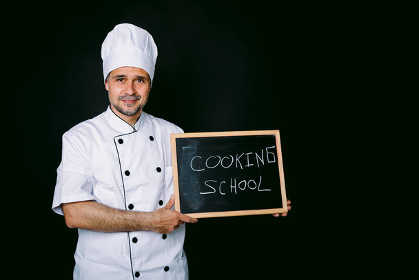 白い帽子とジャケットに身を包んだ料理人は、以下を読む黒板を持っています:黒い背景に調理学校。レストラン、料理、調理学校のコンセプト. - 写真・画像