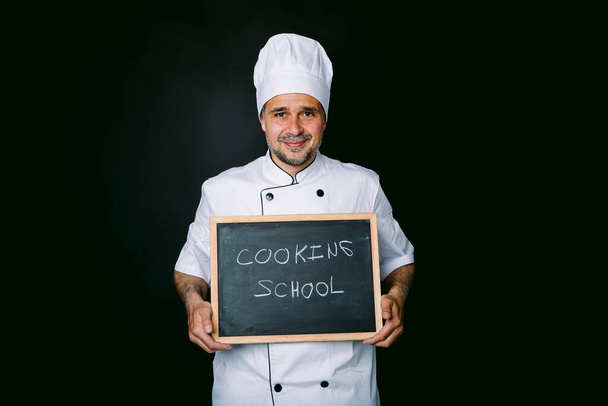 Cook vêtu d'un chapeau et d'une veste blancs tient un tableau noir qui se lit : Cooking School sur un fond noir. Restaurant, cuisine et concept d'école de cuisine. - Photo, image
