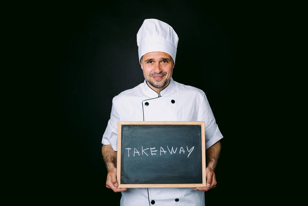 Szakács öltözött fehér kabát és kalap kezében egy táblán, hogy ez áll: "Vigye el" egy fekete háttér. Éttermi és elviteli koncepció. - Fotó, kép