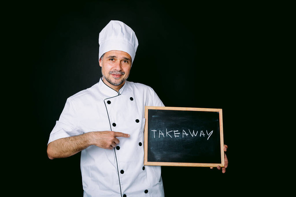 Szakács öltözött fehér kabát és kalap kezében egy táblán, hogy ez áll: "Vigye el" egy fekete háttér. Éttermi és elviteli koncepció. - Fotó, kép
