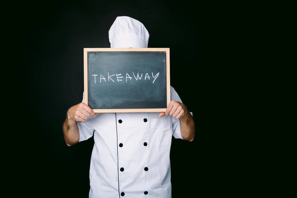 白いジャケットと帽子に身を包んだ料理人は、黒の背景に「離陸」と書かれた黒板を持っています。レストランとテイクアウトのコンセプト. - 写真・画像