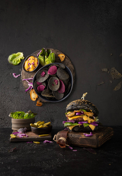 Black burger with cheese,mushrooms,beef cutlet,sauce,greenery and black crisps on dark background.Trend photo in two planes. - Foto, immagini