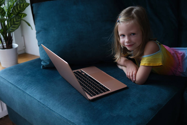 Маленькая улыбающаяся девочка лежит на диване с ноутбуком крупным планом - Фото, изображение