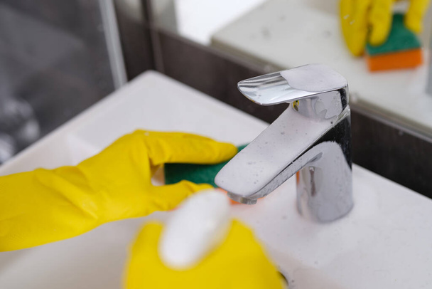 Person in gelben Handschuhen wäscht Waschbecken mit Waschlappen und sprüht Putzschaum auf Wasserhahn - Foto, Bild
