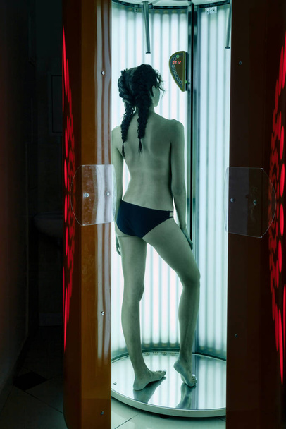 Portrait of cute elegant young woman enjoying tan in modern solarium. Healthy young female in vertical solarium, gets pleasure and enjoys. Concept of healthy lifestyle and beautiful well-groomed skin - 写真・画像