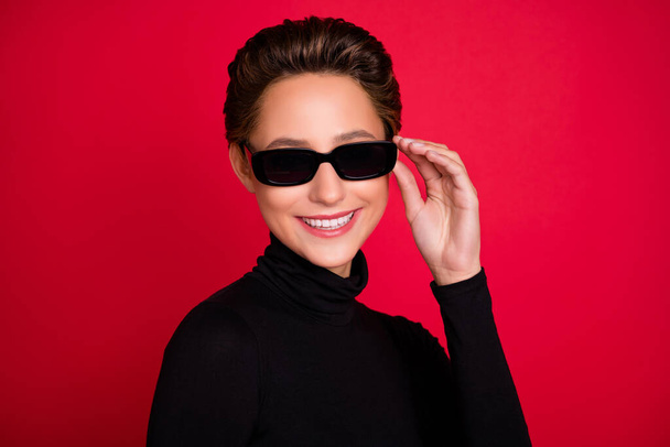 Photo of young woman happy positive smile hand touch sunglass cool isolated over red color background - Fotografie, Obrázek