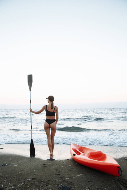 beautiful sports girl in a black swimsuit goes with an orange kayak to swim in the sea at sunset - Foto, Imagem
