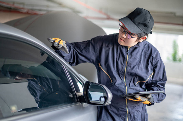 Азиатский автомеханик держит цифровой планшет, проверяя зеркало крыла автомобиля. Инженер по техническому обслуживанию, работающий в автомобильной промышленности. Концепция технического обслуживания и ремонта автомобилей - Фото, изображение