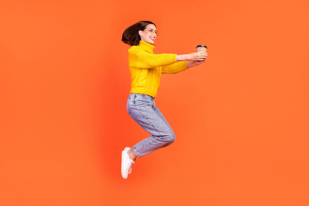 Full body profile side photo of young woman happy positive smile give cup of coffee jump up isolated over orange color background - Фото, зображення