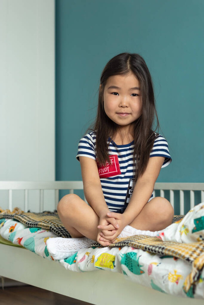 Adorable little asian girl child with long hair sitting in lotus position on bed in nursery at home - 写真・画像