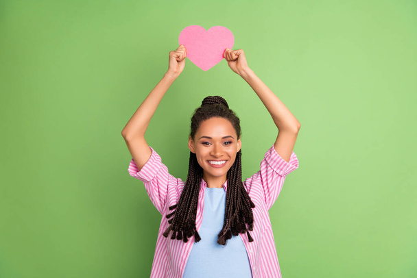 Photo of dreamy shiny dark skin woman dressed striped shirt rising pink heart smiling isolated green color background - Фото, изображение